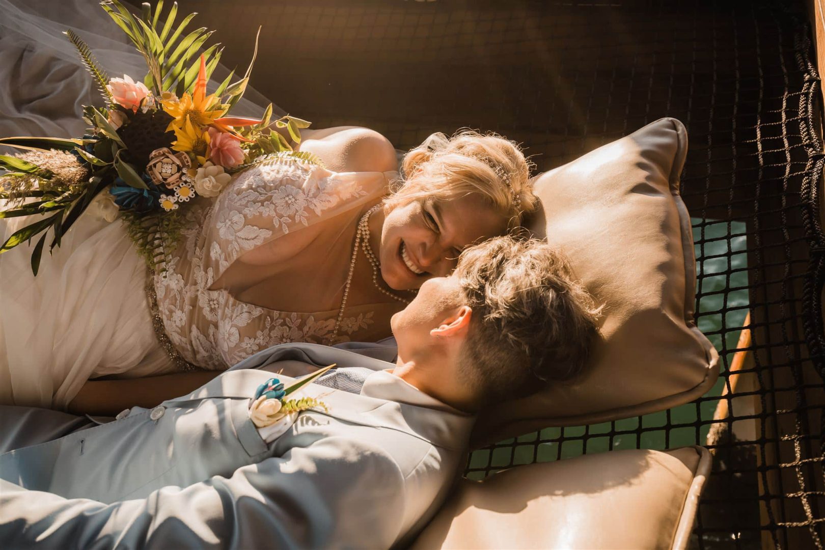 A couple in wedding attire lying in the rope hammock smiling and laughing